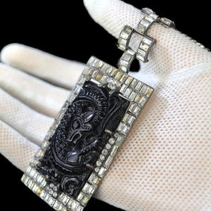 May include: A black and silver pendant with a rectangular black stone carved with a floral design. The stone is surrounded by a border of clear rhinestones. The pendant is attached to a chain made of silver links with clear rhinestones.