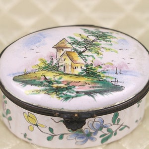 May include: A white porcelain trinket box featuring a hand-painted scene of a small house with a tower, trees, and a river. The lid of the box is decorated with a floral design in shades of blue, yellow, and green.