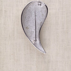 May include: Silver-toned brooch shaped like a teardrop with a textured surface. The brooch has a pin and catch fastening on the back.