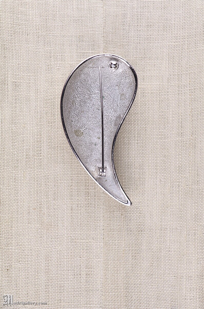 May include: Silver-toned brooch shaped like a teardrop with a textured surface. The brooch has a pin and catch fastening on the back.