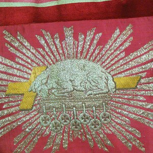 May include: A red fabric with a gold and silver embroidered design. The design features a sunburst with a crown in the center.