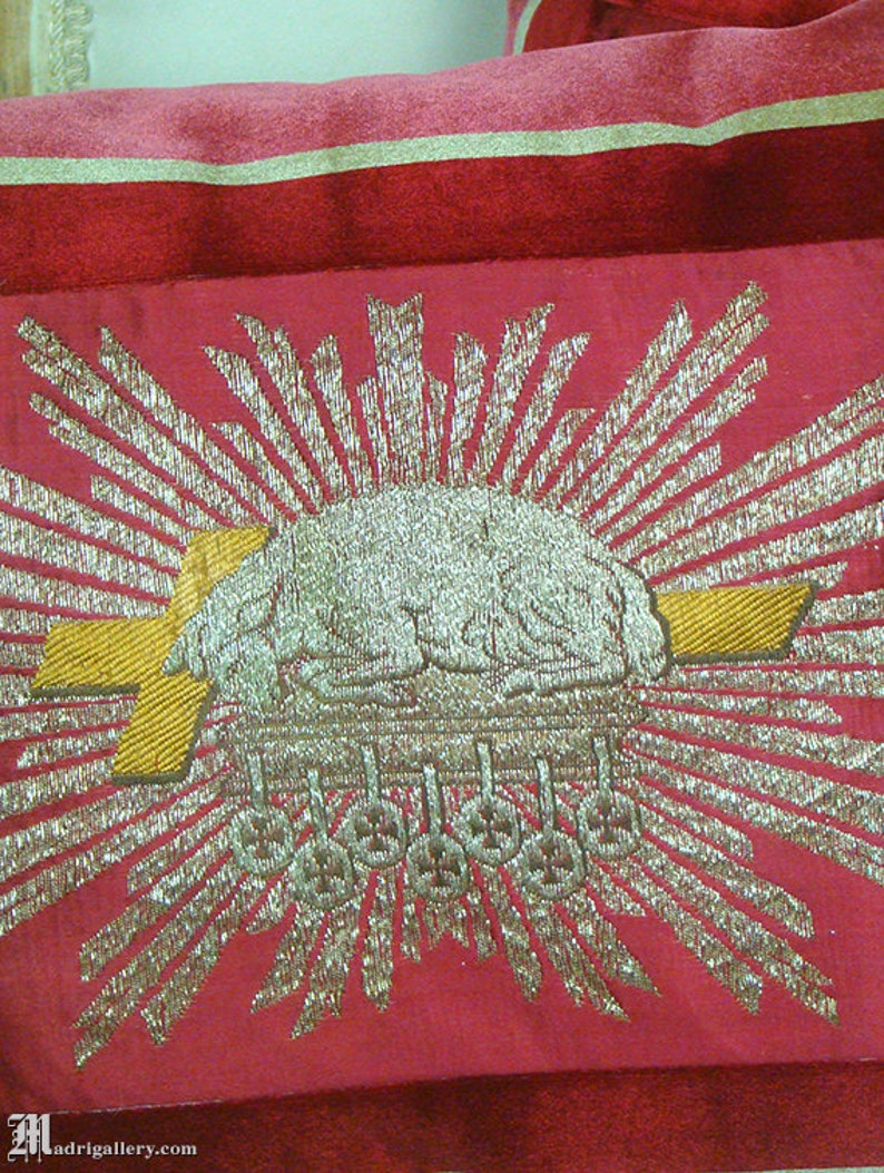 May include: A red fabric with a gold and silver embroidered design. The design features a sunburst with a crown in the center.