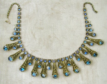 May include: A vintage necklace with a gold tone metal chain and blue iridescent rhinestones. The necklace features a cascading design with teardrop-shaped rhinestones.