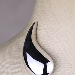 May include: A silver, teardrop-shaped brooch with a smooth, reflective surface.