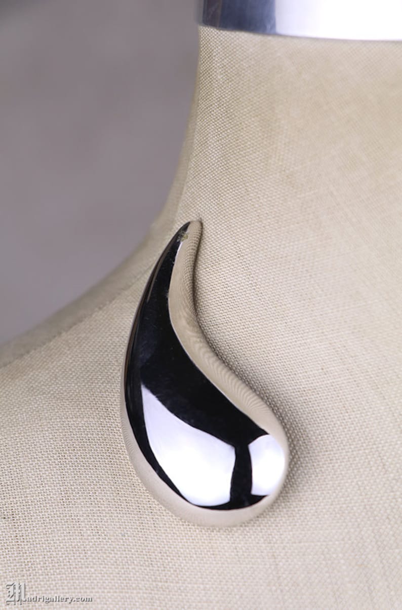 May include: A silver, teardrop-shaped brooch with a smooth, reflective surface.