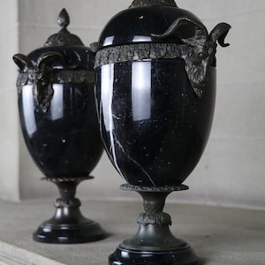 May include: Two black marble urns with bronze accents and ram head finials. The urns are on a stone surface.