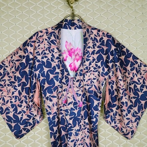 May include: A navy blue kimono with a repeating pink floral pattern. The kimono is hanging on a gold-colored hook. The interior lining is white with a pink floral design. A pink tie with tassels is visible.