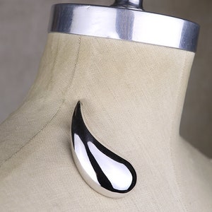 May include: A silver teardrop-shaped pendant brooch, displayed on a fabric-covered mannequin.