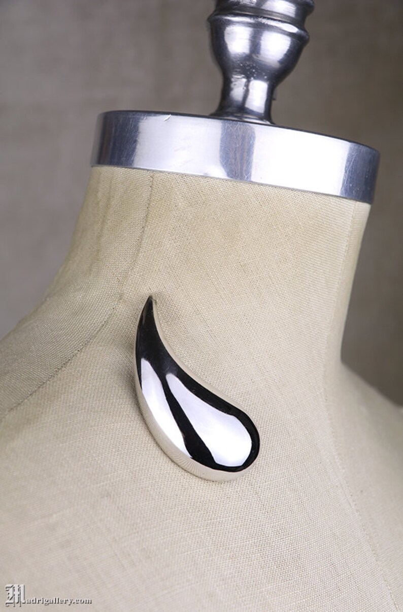 May include: A silver teardrop-shaped pendant brooch, displayed on a fabric-covered mannequin.