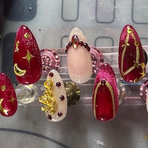 May include: A set of ten artificial nails in various designs. The nails feature a mix of red, white, and pink colors, adorned with gold accents, rhinestones, and celestial motifs like stars and moons. The nails are oval-shaped.