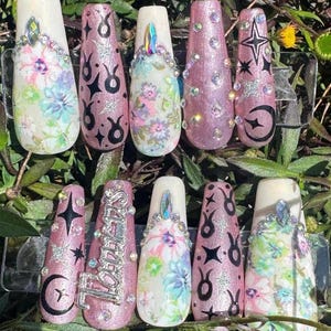 May include: Set of ten long, pink and white press-on nails with various designs. Some nails feature floral patterns, others have black and silver glitter, and some have black stars and moons. One nail has the word "Taurus" written in silver glitter.