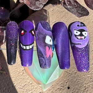 Może przedstawiać: Pięć długich, fioletowych i błyszczących sztucznych paznokci. Trzy paznokcie zdobią postacie z kreskówek z białymi, czerwonymi i różowymi akcentami. Paznokcie są prezentowane na przezroczystym, iryzującym stojaku.