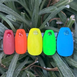 May include: Five fake nails with a rainbow color scheme. The nails are painted in shades of red, orange, yellow, green, and blue. Each nail has a small, sparkly accent.