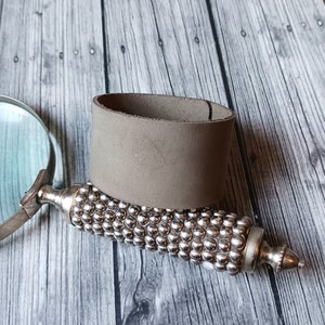 May include: A wide, matte taupe leather cuff bracelet resting on the handle of a magnifying glass. The handle features a textured, silver-toned metallic surface covered in small, raised spherical studs, ending in a pointed, polished metal tip.