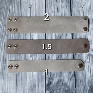 May include: Three rectangular light grey leather wristbands of varying widths, labelled 2, 1.5, and 1. Each band features two silver-toned metal snap fasteners on one end and a single matching snap fastener on the opposite end for closure.