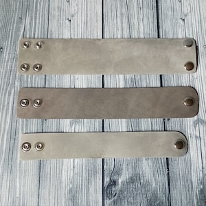 May include: Three rectangular leather bracelets in varying shades of grey and taupe. Each bracelet features two silver-toned metal snap buttons on one end and a single matching snap button on the opposite end for adjustable closure and secure fastening.