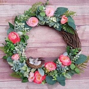 May include: A circular wreath constructed from brown twigs, decorated with pink ranunculus flowers, various green eucalyptus leaves, and small white floral sprigs. A small woven nest containing two grey birds and a white egg sits at the bottom.