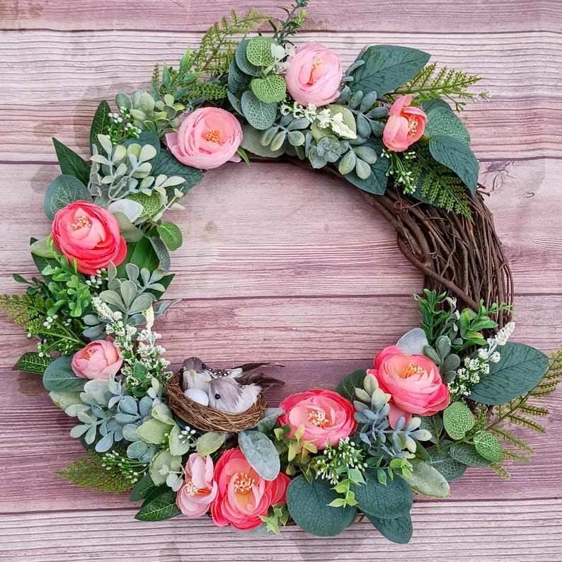May include: A circular wreath constructed from brown twigs, decorated with pink ranunculus flowers, various green eucalyptus leaves, and small white floral sprigs. A small woven nest containing two grey birds and a white egg sits at the bottom.
