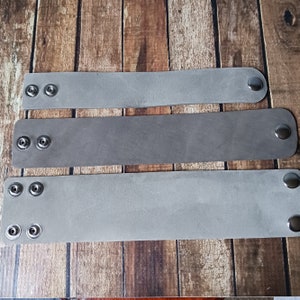 May include: Three grey leather bracelets of varying widths arranged horizontally. Each bracelet features two silver-toned metal snap buttons on one end and a single matching snap button on the opposite end, providing an adjustable closure mechanism.