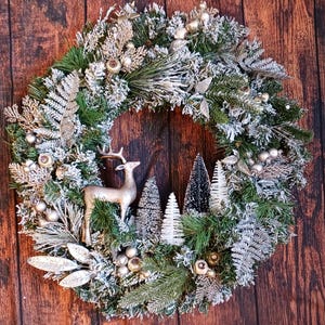 May include: A circular wreath featuring green pine branches with a dusting of white snow. The wreath is decorated with silver fern leaves, small metallic spheres, a silver-toned deer figurine, and several miniature bottle brush trees.