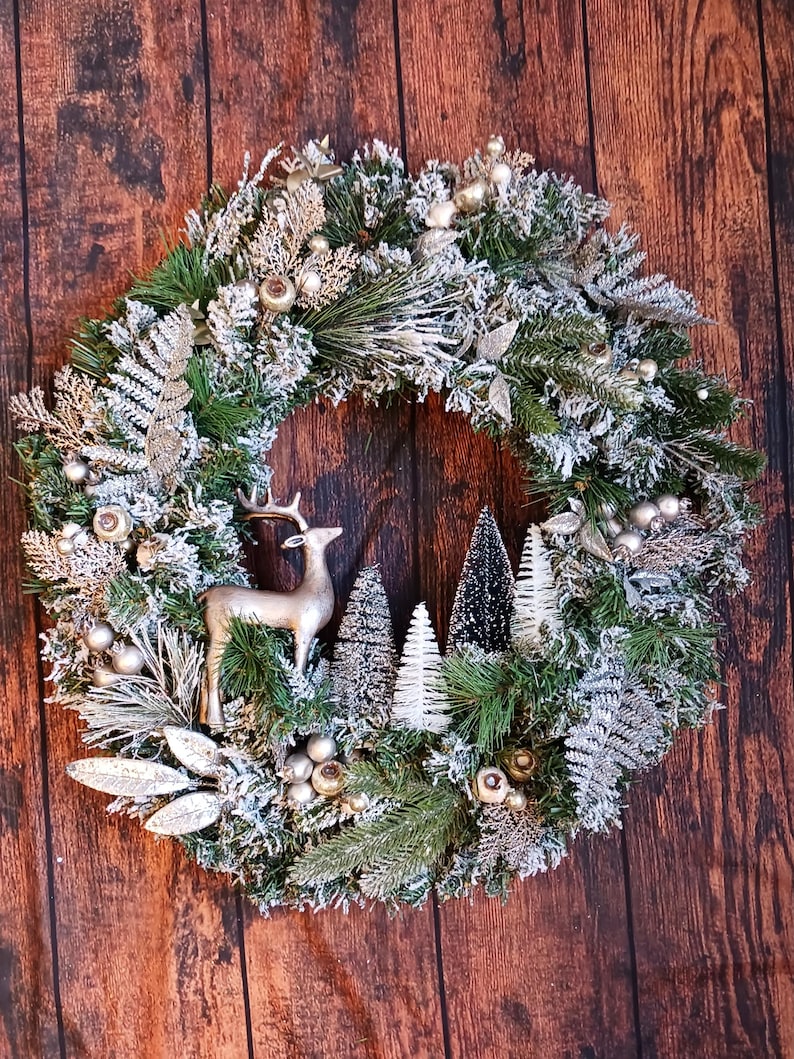 May include: A circular wreath featuring green pine branches with a dusting of white snow. The wreath is decorated with silver fern leaves, small metallic spheres, a silver-toned deer figurine, and several miniature bottle brush trees.