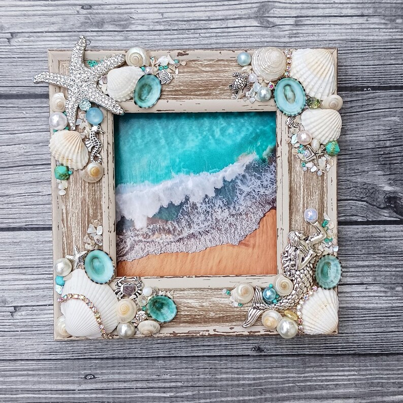 May include: A square picture frame with a distressed light wood finish, decorated with white seashells, pearls, turquoise stones, a silver starfish, and a silver mermaid figure. The centre displays an aerial view of turquoise ocean waves.