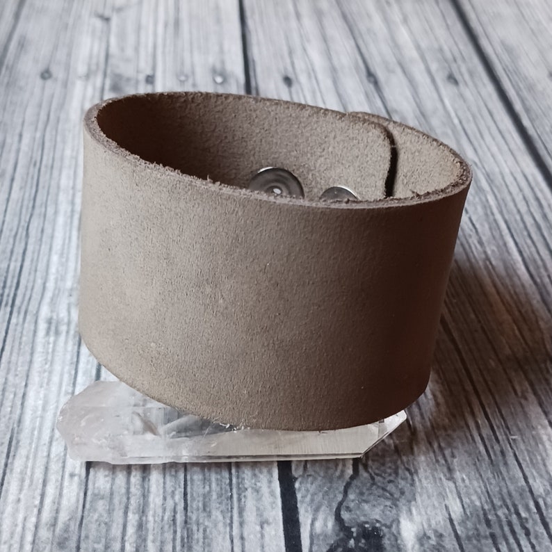 May include: A wide, light brown leather cuff bracelet with a smooth, matte texture. The bracelet features two silver-toned metal snap fasteners on the interior side, allowing for an adjustable closure. The leather band is thick and solid in colour.