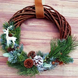 May include: A rustic Christmas wreath made of woven brown branches, decorated with green pine needles, pine cones, and a white deer figurine. The wreath is accented with frosted greenery and a brown ribbon for hanging.
