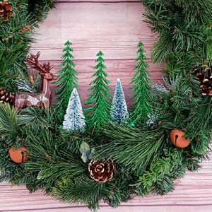May include: A circular holiday wreath made of green pine needles featuring a brown geometric deer figure, three glittery green bottle brush trees, two smaller white bottle brush trees, brown pinecones, and two round copper-coloured jingle bells.