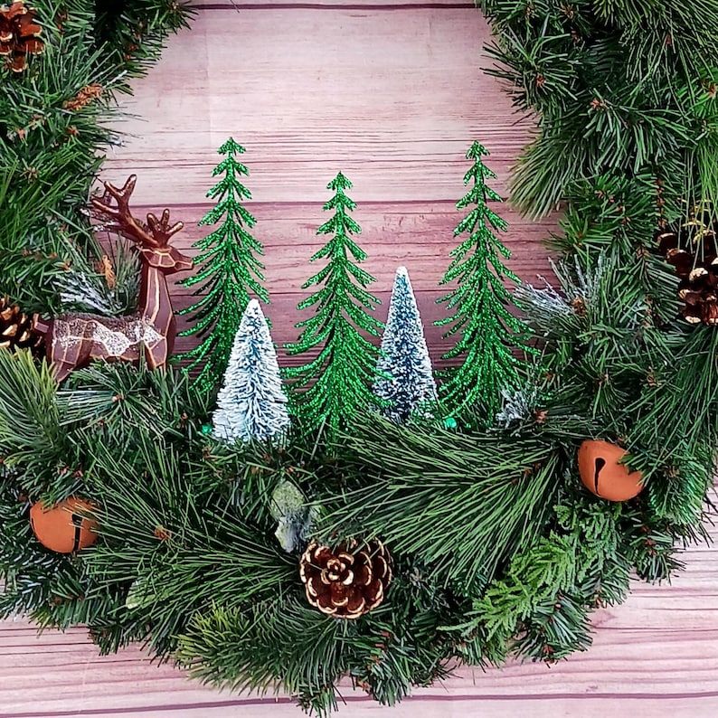 May include: A circular holiday wreath made of green pine needles featuring a brown geometric deer figure, three glittery green bottle brush trees, two smaller white bottle brush trees, brown pinecones, and two round copper-coloured jingle bells.