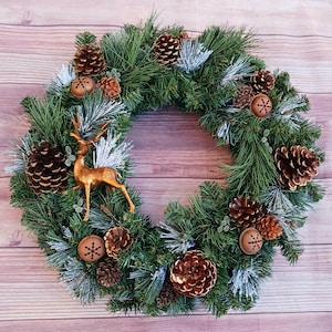May include: A circular wreath made of green pine needles with white frosted tips. The wreath is decorated with brown pinecones of various sizes, small metallic copper-coloured jingle bells with snowflake cutouts, and a central gold-toned deer figure.