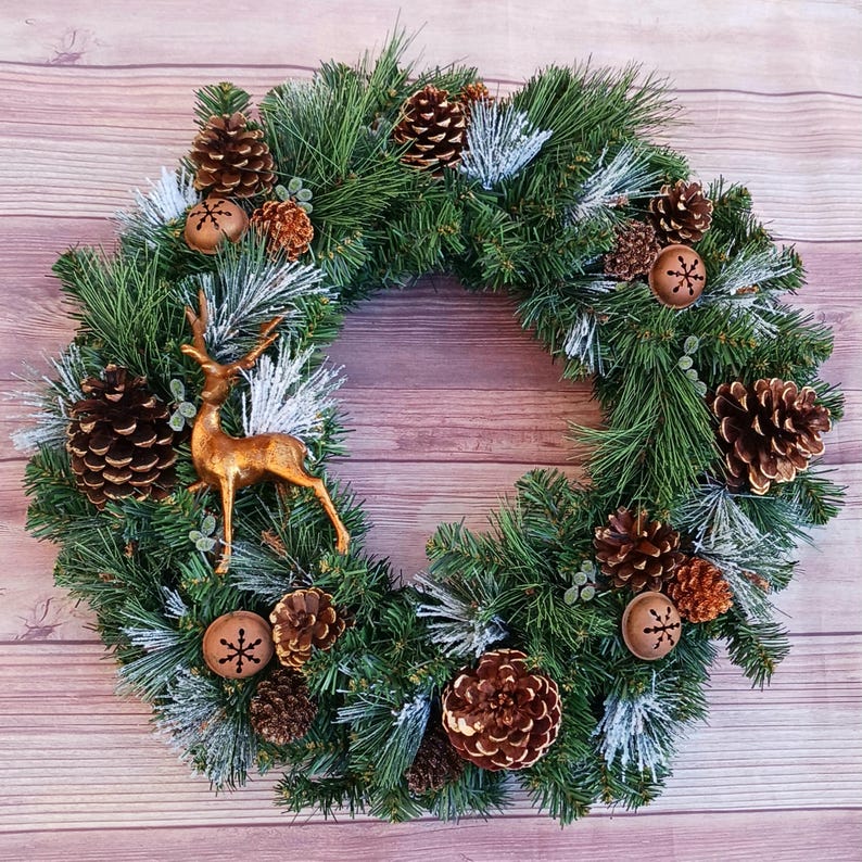 May include: A circular wreath made of green pine needles with white frosted tips. The wreath is decorated with brown pinecones of various sizes, small metallic copper-coloured jingle bells with snowflake cutouts, and a central gold-toned deer figure.