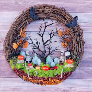May include: A round grapevine wreath featuring a graveyard scene with two plastic skeletons, miniature grey tombstones, and a central black glittery tree. The base is covered in green moss, orange berries, and small pumpkins, with two black bats attached.