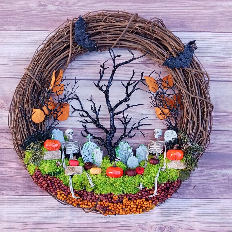 May include: A round grapevine wreath featuring a graveyard scene with two plastic skeletons, miniature grey tombstones, and a central black glittery tree. The base is covered in green moss, orange berries, and small pumpkins, with two black bats attached.
