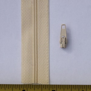 May include: A close-up of a beige zipper tape with a plastic zipper pull. The tape is measured with a yellow tape measure that reads "DURA NY-Coat™ 4 5 NYLON COAT 6 3 9 101 2 3 4 5".