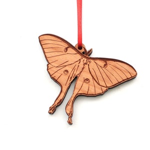 May include: Wooden ornament shaped like a luna moth with a red ribbon attached. The ornament is a light brown color and has a detailed design of the moth's wings and body.