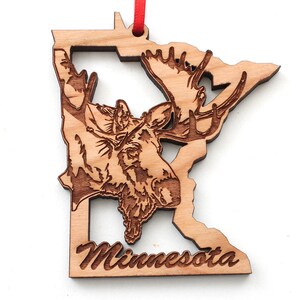 May include: Wooden ornament shaped like the state of Minnesota with a moose head carved into the outline. The ornament is painted brown and has the word "Minnesota" written on the bottom.