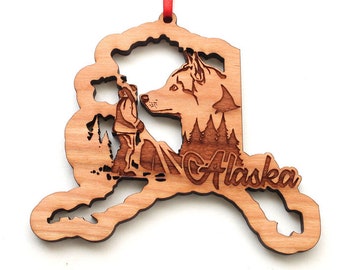 May include: Wooden ornament shaped like the state of Alaska with a husky dog and a person standing in the mountains. The word "Alaska" is engraved on the ornament.