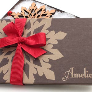 Op de afbeelding: Een bruine cadeaudoos met een sneeuwvlokkenpatroon en een rood lint. De doos is gepersonaliseerd met de naam "Amelia".