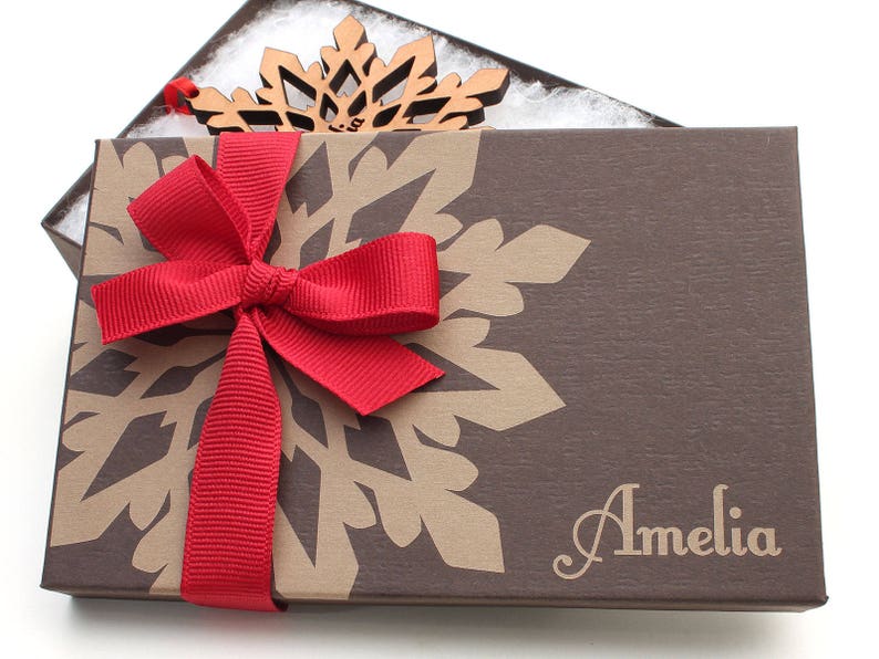 Op de afbeelding: Een bruine cadeaudoos met een sneeuwvlokkenpatroon en een rood lint. De doos is gepersonaliseerd met de naam "Amelia".