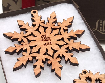 Op de afbeelding: Een houten sneeuwvlok ornament met de naam "Lily" en het jaar "2016" erop gegraveerd. Het ornament zit in een witte doos met een bruin deksel.