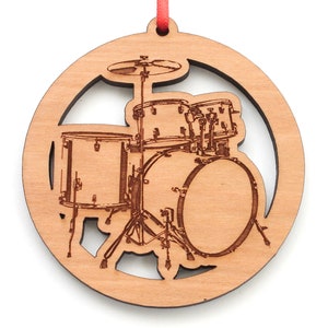 May include: A wooden Christmas ornament shaped like a circle with a cutout design of a drum set. The ornament is made of light brown wood and has a red ribbon attached to the top.