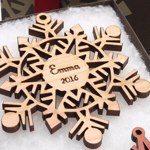 Op de afbeelding: Een houten sneeuwvlok ornament met een gepersonaliseerde inscriptie die "Emma 2016" luidt. Het ornament is lichtbruin en heeft een delicaat, ingewikkeld ontwerp.