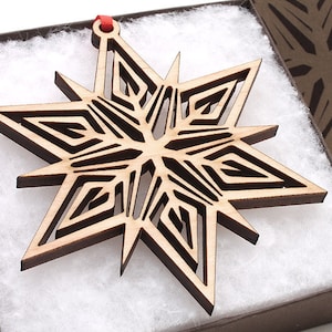 May include: A wooden snowflake ornament with a detailed, intricate design. The ornament is light brown in color and has a red string attached to the top.