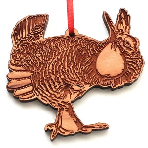 May include: A wooden ornament of a Greater Prairie Chicken, a bird with a brown and white patterned body, a long neck, and a small head. The ornament is carved in a detailed style and has a red ribbon attached to the top.