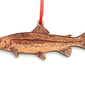 May include: A wooden ornament shaped like a trout fish with a red ribbon attached. The ornament is detailed with a woodgrain pattern and a realistic depiction of the fish's scales.