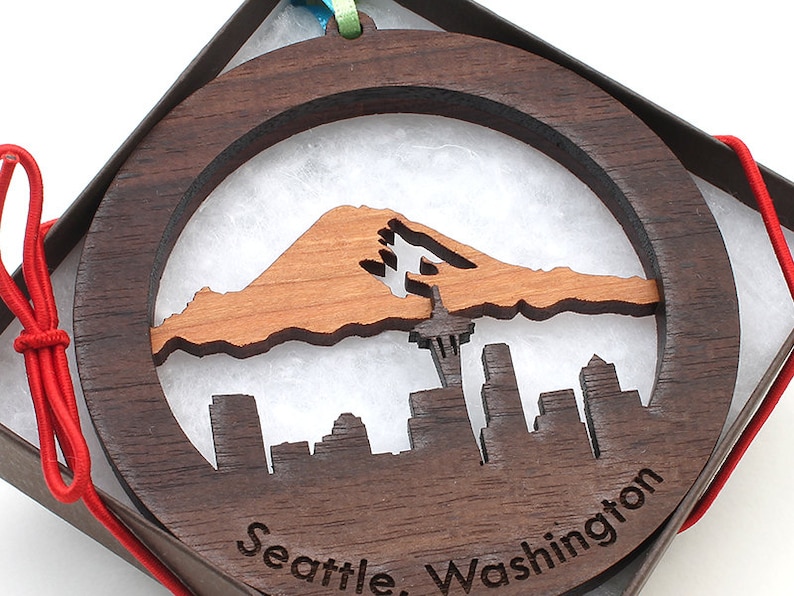May include: A round wooden ornament with a cutout design of the Seattle skyline and Mount Rainier. The ornament is brown with a natural wood finish and has the text "Seattle, Washington" engraved on the bottom.