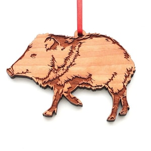 May include: A wooden ornament shaped like a brown and white collared peccary, also known as a javelina, walking to the right. The ornament is made of wood and has a red ribbon attached to the top.