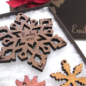 Op de afbeelding: Een set van drie houten sneeuwvlokversieringen in een bruine doos. De grootste sneeuwvlok is gepersonaliseerd met de naam "Emily" en het jaar "2015".