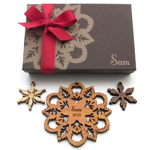 Op de afbeelding: Een set van drie houten sneeuwvlokversieringen in een bruine cadeaudoos met een rood lint. De doos is versierd met een bruin en gouden bloemenpatroon en de naam "Sam" staat op de voorkant gedrukt. De grootste sneeuwvlokversiering heeft de naam "Sam" en het jaar "2015" erop gegraveerd.
